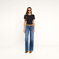 Woman wearing sunglasses, a relaxed fit black Aileen Cotton Crewneck Tee crafted from soft Supima cotton, blue flared jeans with a brown belt, and brown heeled boots, standing against a plain beige background.