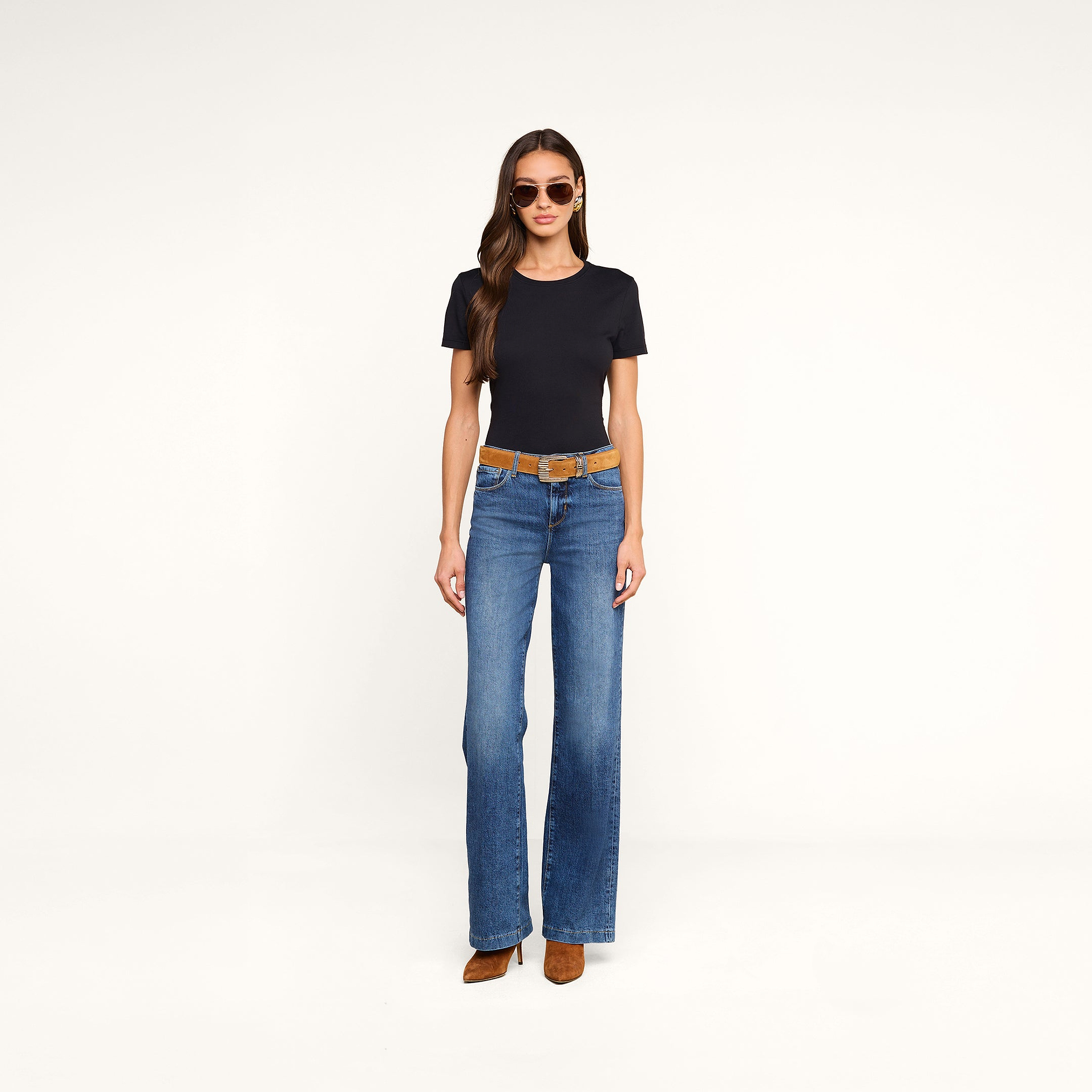 Woman wearing sunglasses, a relaxed fit black Aileen Cotton Crewneck Tee crafted from soft Supima cotton, blue flared jeans with a brown belt, and brown heeled boots, standing against a plain beige background.