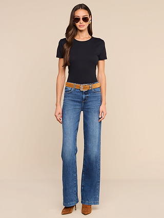 Woman wearing sunglasses, a relaxed fit black Aileen Cotton Crewneck Tee crafted from soft Supima cotton, blue flared jeans with a brown belt, and brown heeled boots, standing against a plain beige background.
