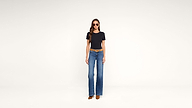 A woman wearing sunglasses, a black t-shirt, blue wide-leg jeans, a brown belt, and brown shoes stands against a plain white background.