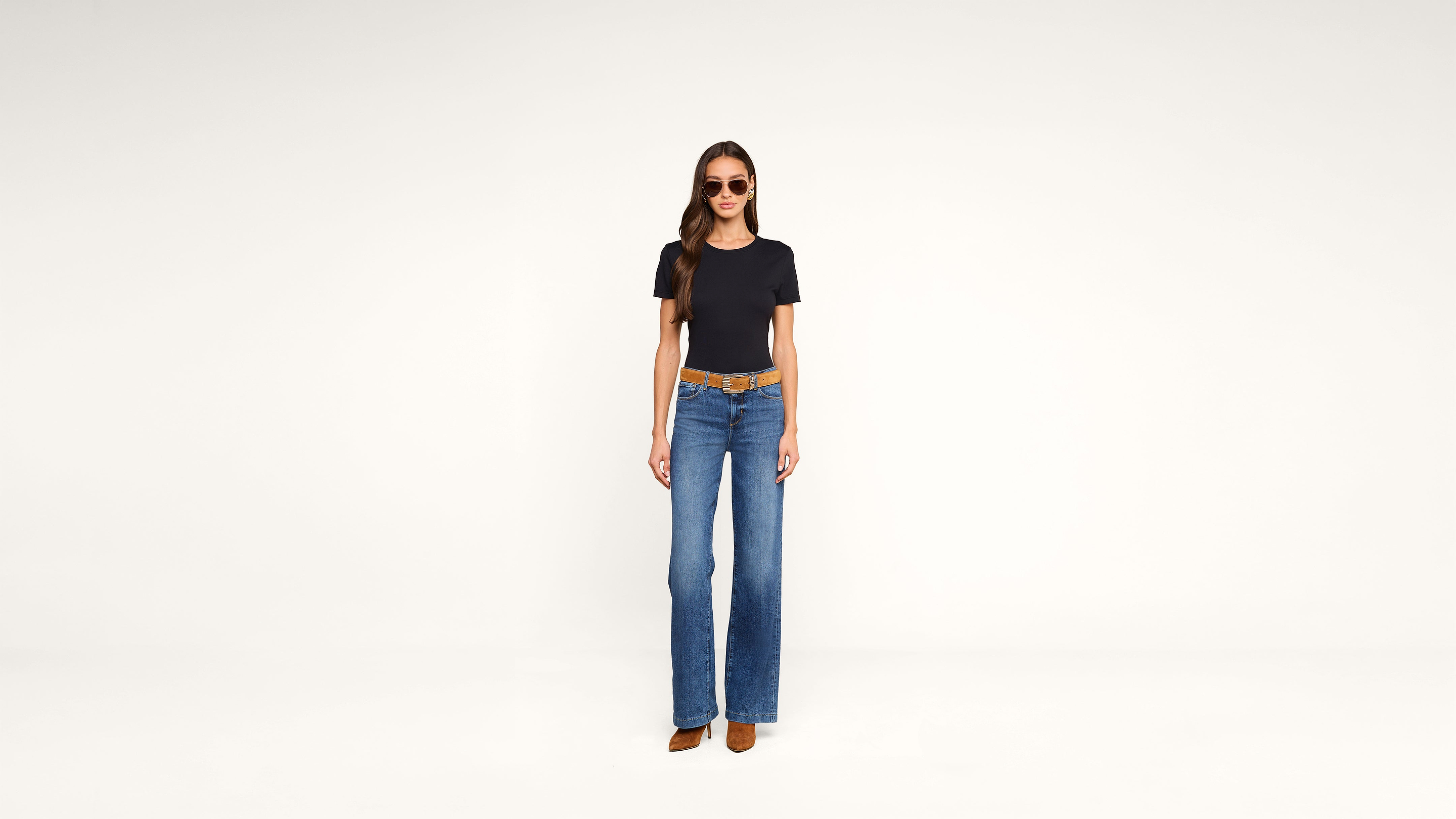A woman wearing sunglasses, a black t-shirt, blue wide-leg jeans, a brown belt, and brown shoes stands against a plain white background.
