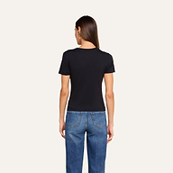 A person with long brown hair, wearing a black Aileen Cotton Crewneck Tee and blue jeans, stands facing away from the camera in front of a plain beige background.