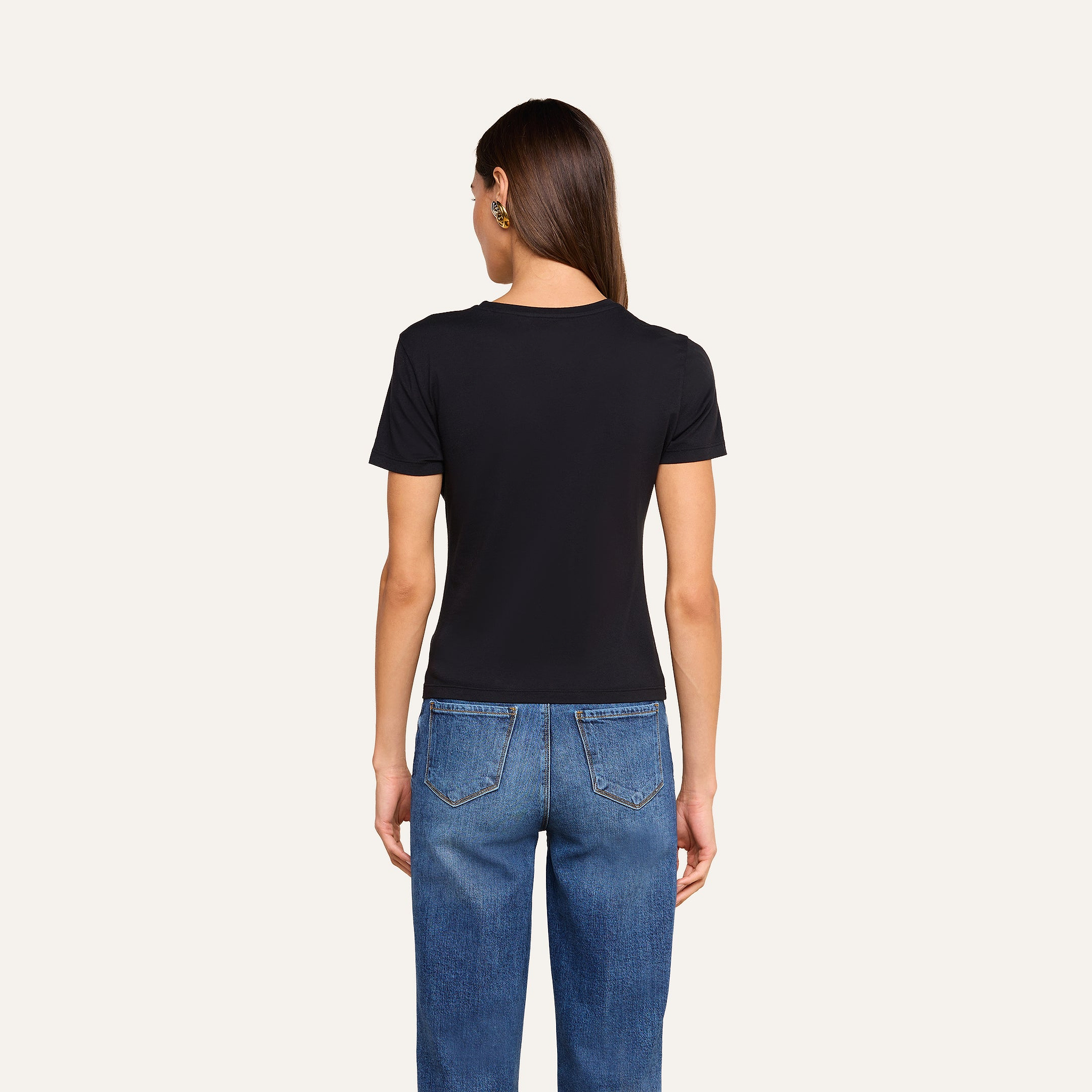 A person with long brown hair, wearing a black Aileen Cotton Crewneck Tee and blue jeans, stands facing away from the camera in front of a plain beige background.