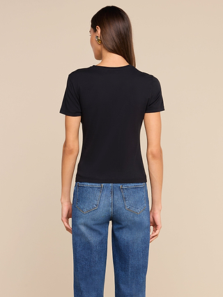 A person with long brown hair, wearing a black Aileen Cotton Crewneck Tee and blue jeans, stands facing away from the camera in front of a plain beige background.