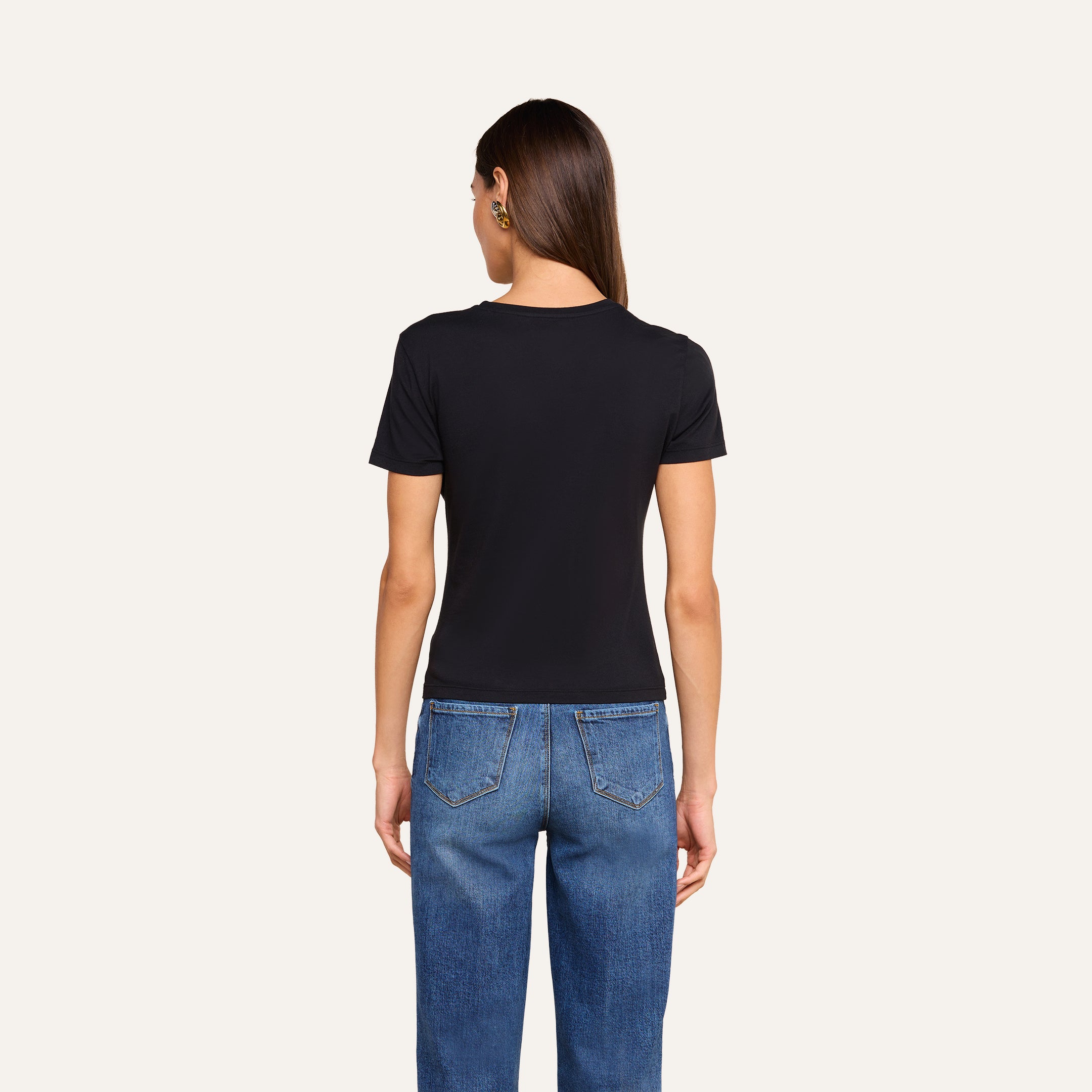 A person with long brown hair, wearing a black Aileen Cotton Crewneck Tee and blue jeans, stands facing away from the camera in front of a plain beige background.