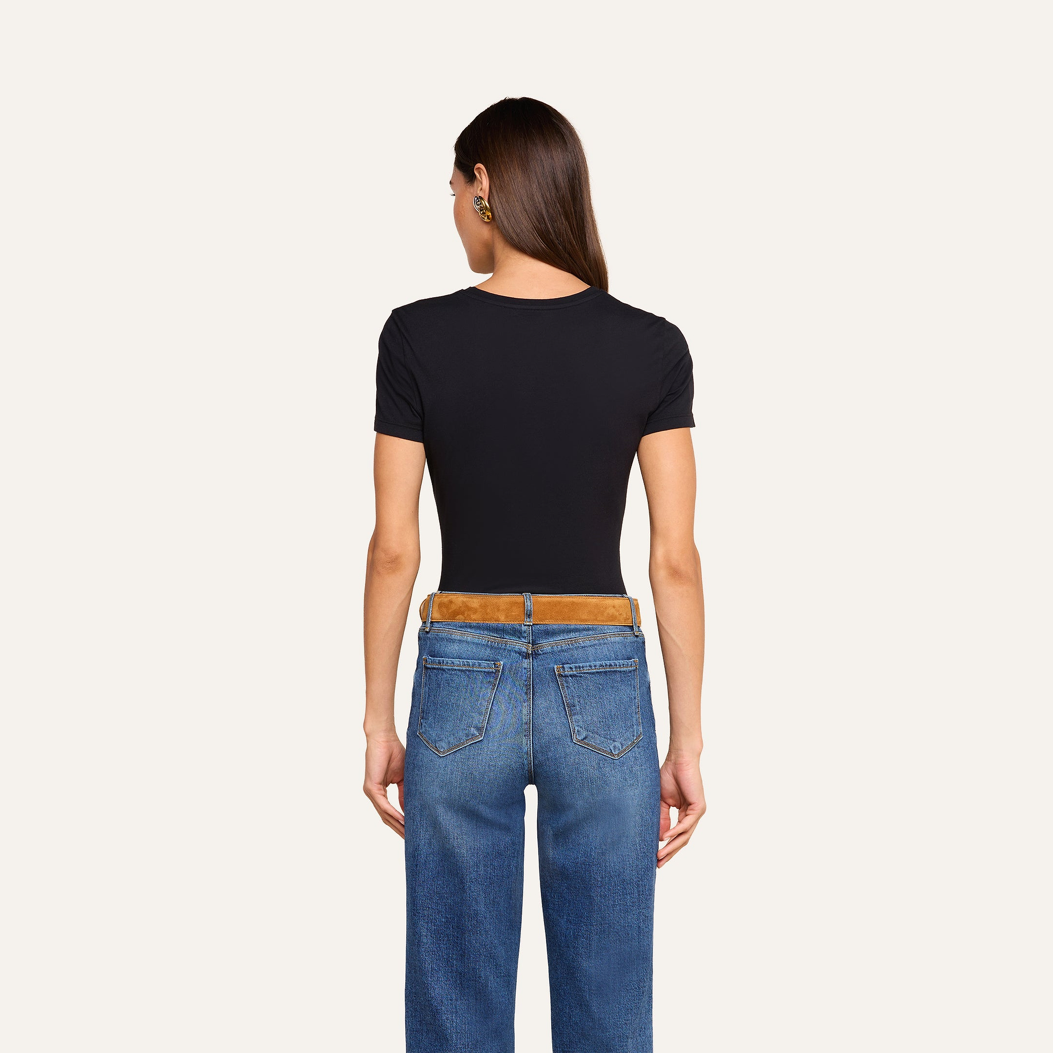 A woman with long brown hair stands facing away, wearing the Aileen Cotton Crewneck Tee in black—a relaxed fit crewneck made from Supima cotton—paired with blue jeans and gold earrings, against a plain beige background.