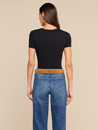 A woman with long brown hair stands facing away, wearing the Aileen Cotton Crewneck Tee in black—a relaxed fit crewneck made from Supima cotton—paired with blue jeans and gold earrings, against a plain beige background.