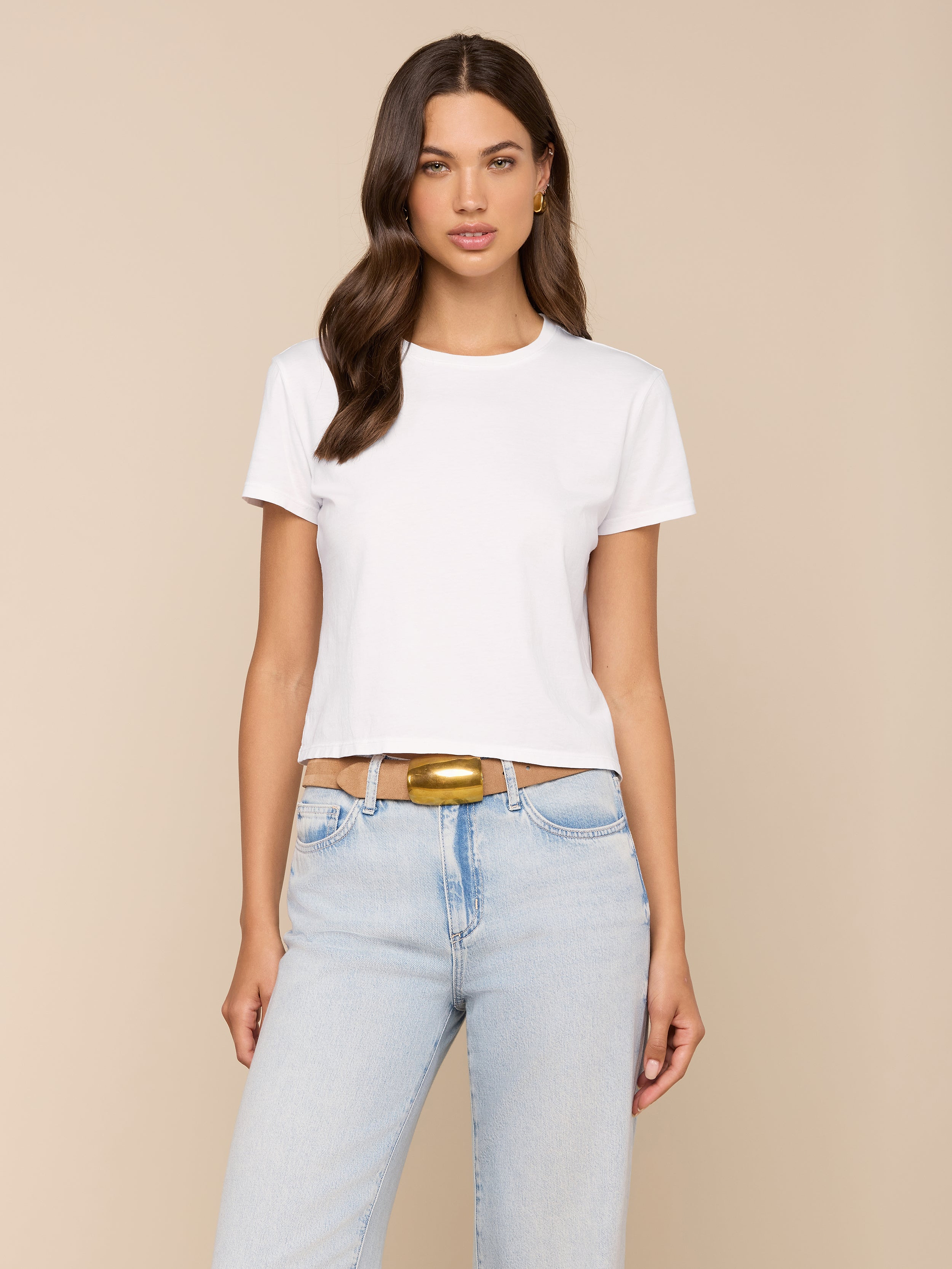 A woman with long brown hair wears the Aileen Cotton Crewneck Tee in plain white, paired with light blue jeans and a tan belt, standing against a plain light background.