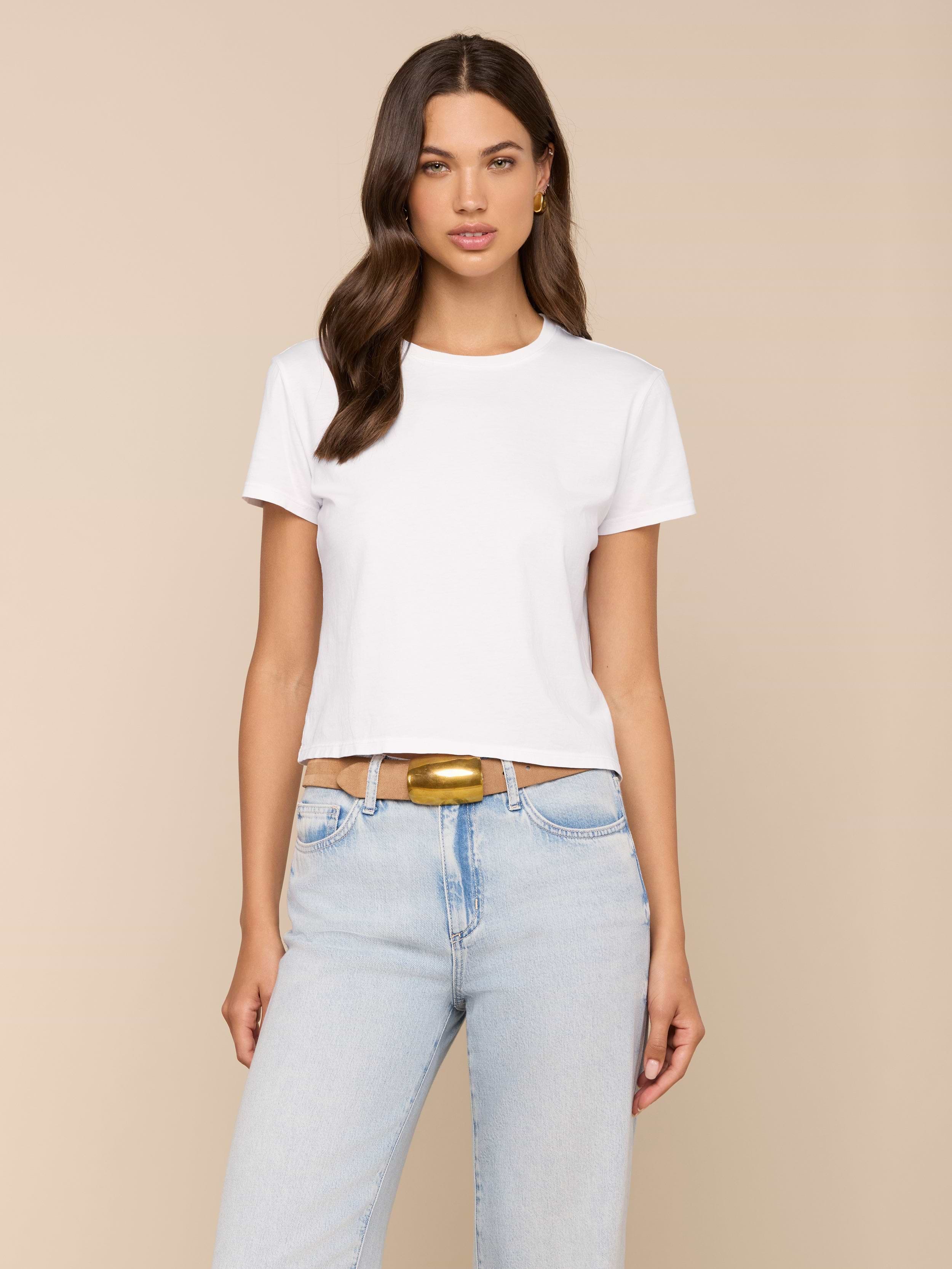 A woman with long brown hair wears the Aileen Cotton Crewneck Tee in plain white, paired with light blue jeans and a tan belt, standing against a plain light background.