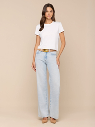 A woman stands against a plain background, wearing the Aileen Cotton Crewneck Tee in white, paired with light blue wide-leg jeans with a brown belt and tan heels. She has long brown hair and a neutral expression.