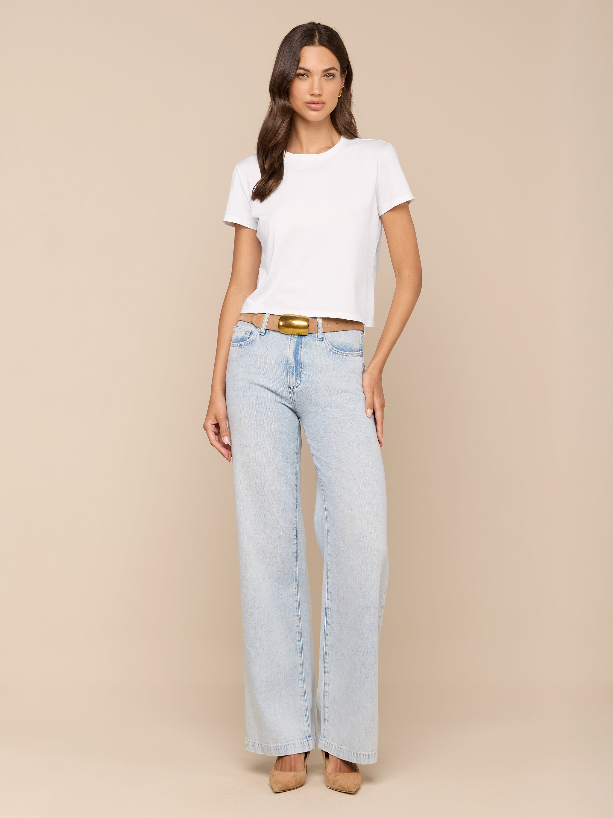 A woman stands against a plain background, wearing the Aileen Cotton Crewneck Tee in white, paired with light blue wide-leg jeans with a brown belt and tan heels. She has long brown hair and a neutral expression.