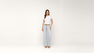 Woman stands against a plain white background wearing a white t-shirt, light blue wide-leg jeans, a brown belt, and beige shoes.