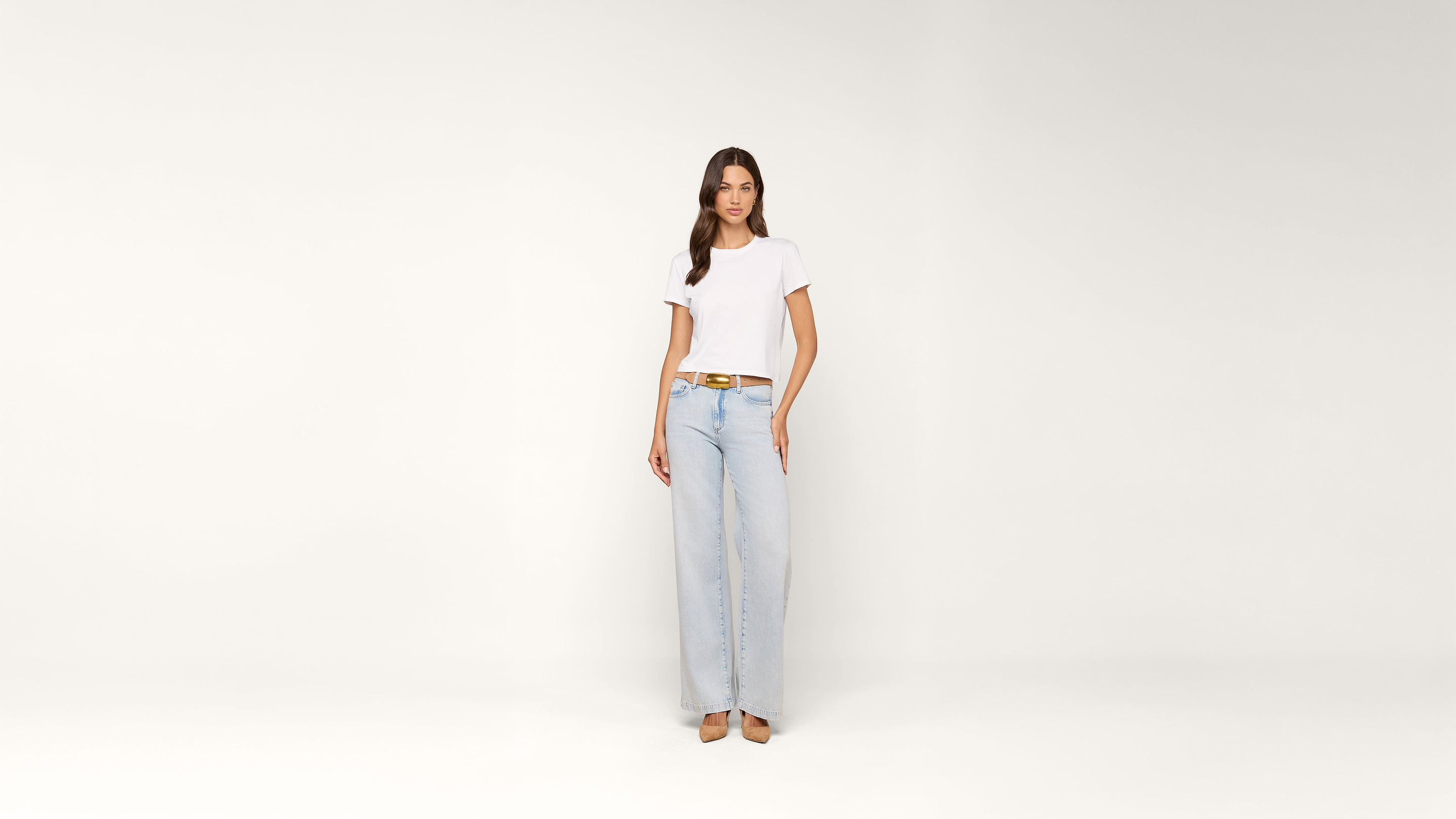 Woman stands against a plain white background wearing a white t-shirt, light blue wide-leg jeans, a brown belt, and beige shoes.