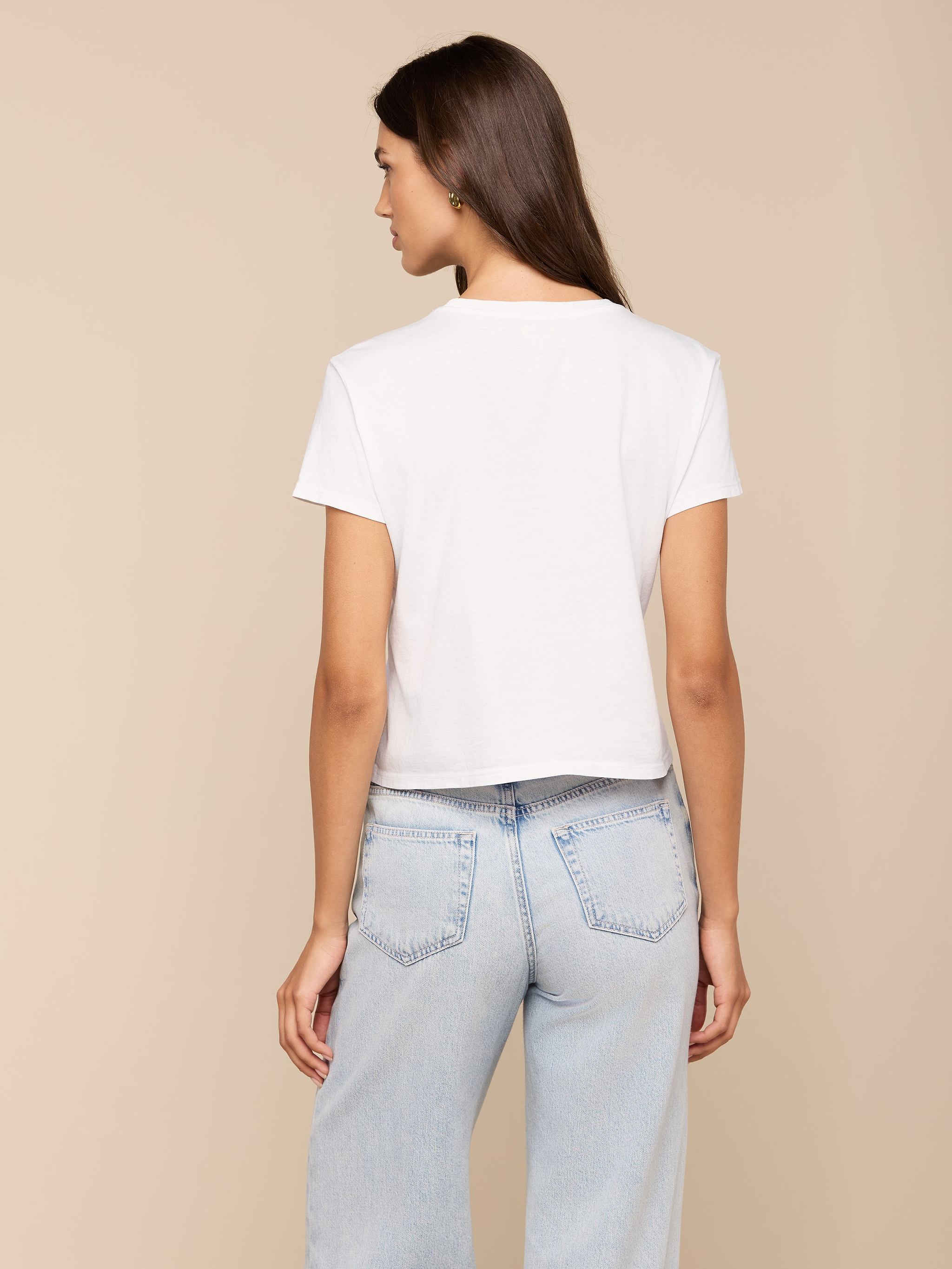 A woman with long brown hair stands with her back to the camera, wearing a white Aileen Cotton Crewneck Tee and light blue jeans against a plain light background.