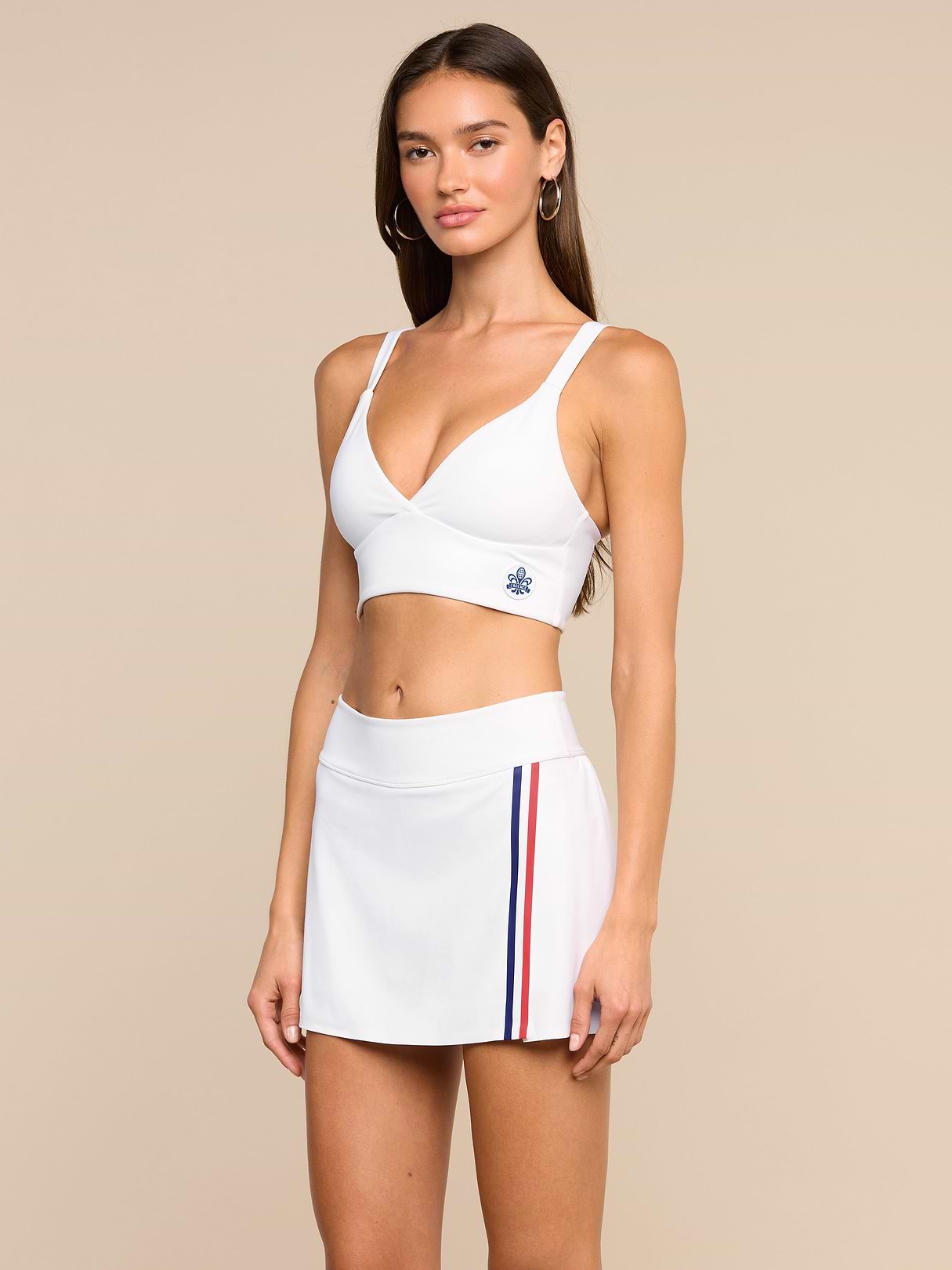 A woman wears the Steffi Tennis Bralette, a white crop top styled as a tennis bralette, paired with a white skirt featuring red and blue side stripes, standing against a plain background while looking toward the camera.