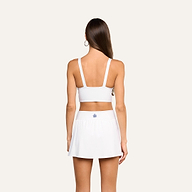 A woman with long brown hair, wearing the Steffi Tennis Bralette in white and a matching white skirt, stands facing away from the camera on a plain background.