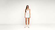 Woman wearing sunglasses, a white athletic tank top, and a white tennis skirt with green accents, standing against a plain white background. She is also wearing white sneakers and socks.