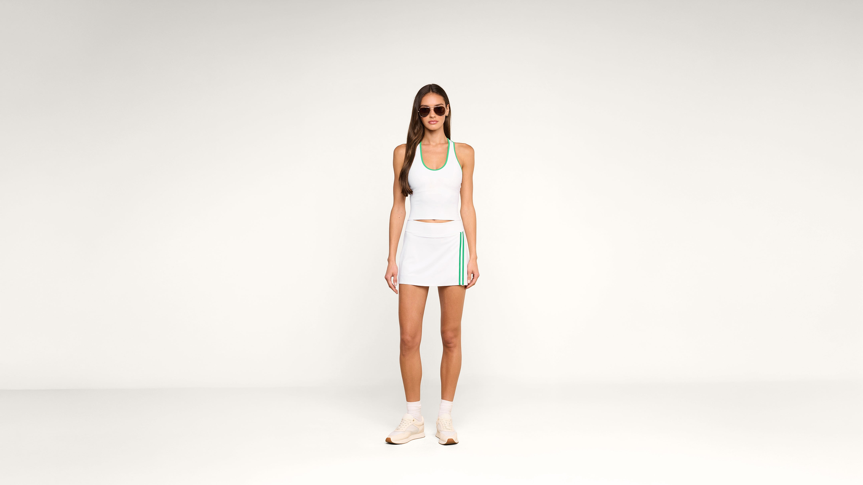 Woman wearing sunglasses, a white athletic tank top, and a white tennis skirt with green accents, standing against a plain white background. She is also wearing white sneakers and socks.