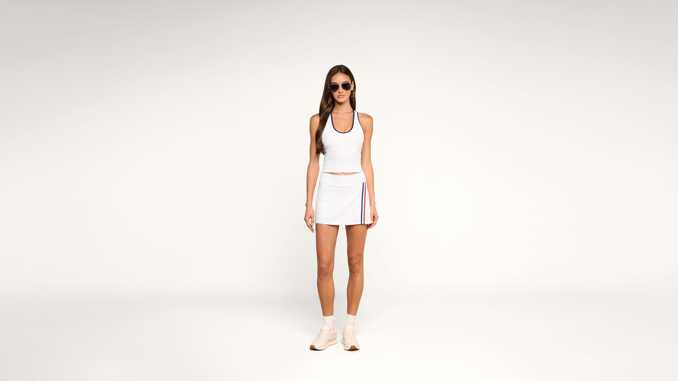 Woman in sunglasses, white tank top, and white skirt with colored stripes stands against a plain white background. She wears white socks and sneakers, with her arms relaxed by her sides.