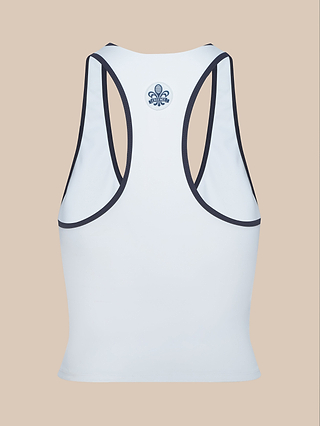 The image features the Martina Racerback Tennis Tank in white with black trim, displayed from the back. Made from performance fabric, it showcases a small blue fleur-de-lis symbol centered near the neckline. The background is beige.