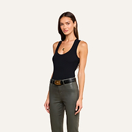 Woman standing against a beige background, wearing the Shania Racerback Tank in black, paired with green pants, a black belt with a gold buckle, gold earrings, and a gold necklace. She has long brown hair down and wears a neutral expression.