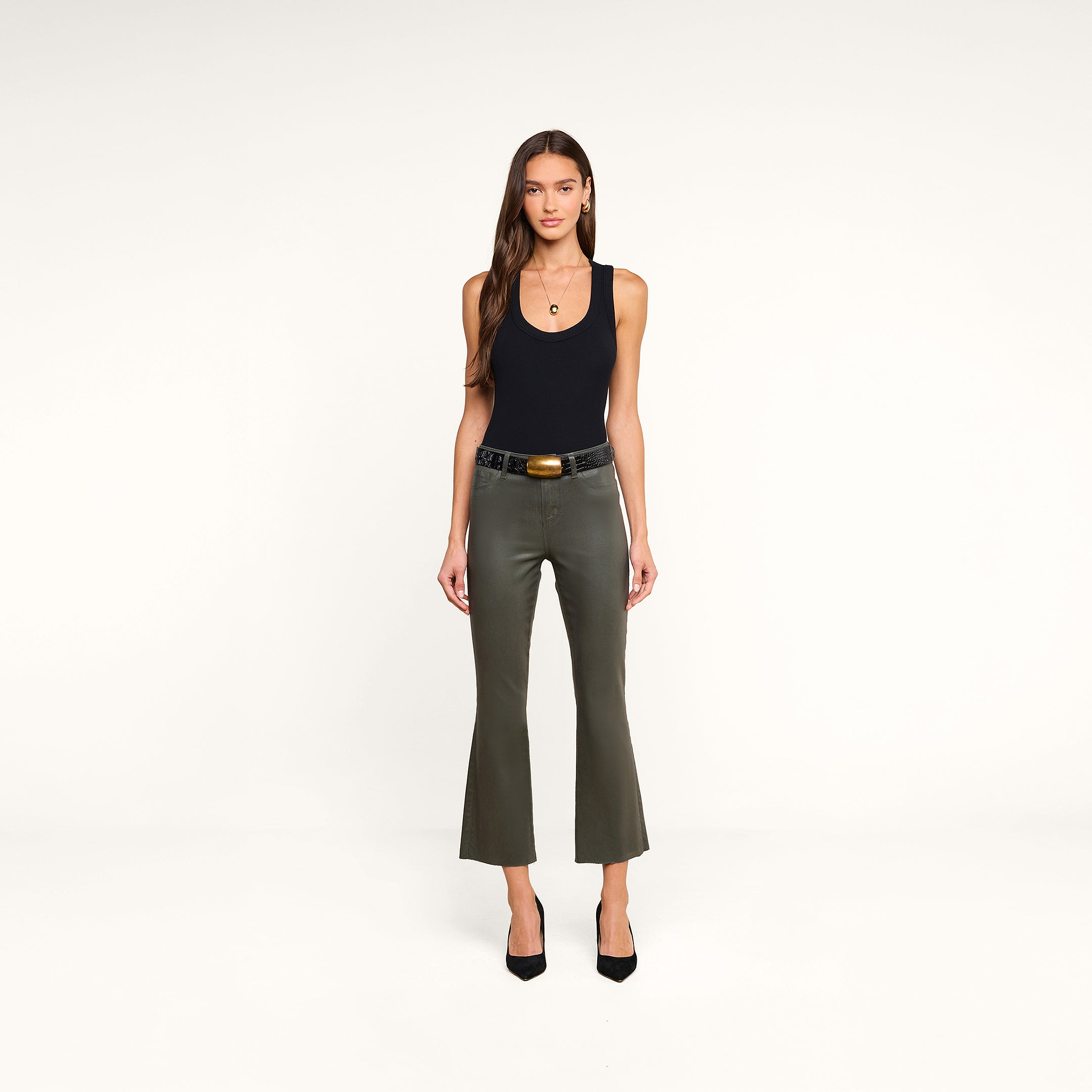 Woman stands facing forward, wearing the Shania Racerback Tank in black, paired with olive-green flared pants, a black belt with a gold buckle, and black heels, against a plain beige background.