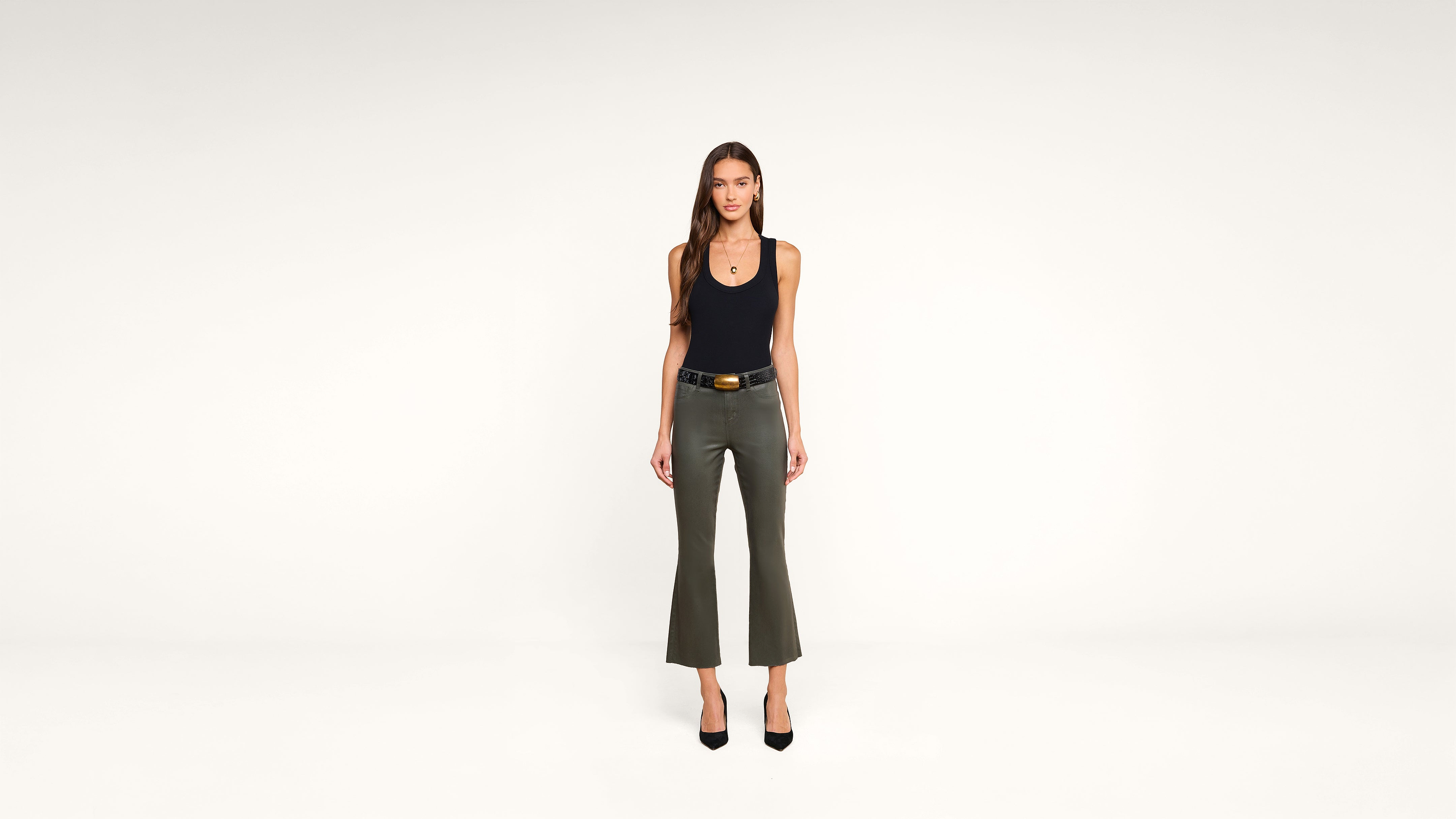 Woman stands facing forward in a minimalist white studio, wearing a black tank top, olive green flared pants, black belt, and black heels. She has long hair and a neutral expression.