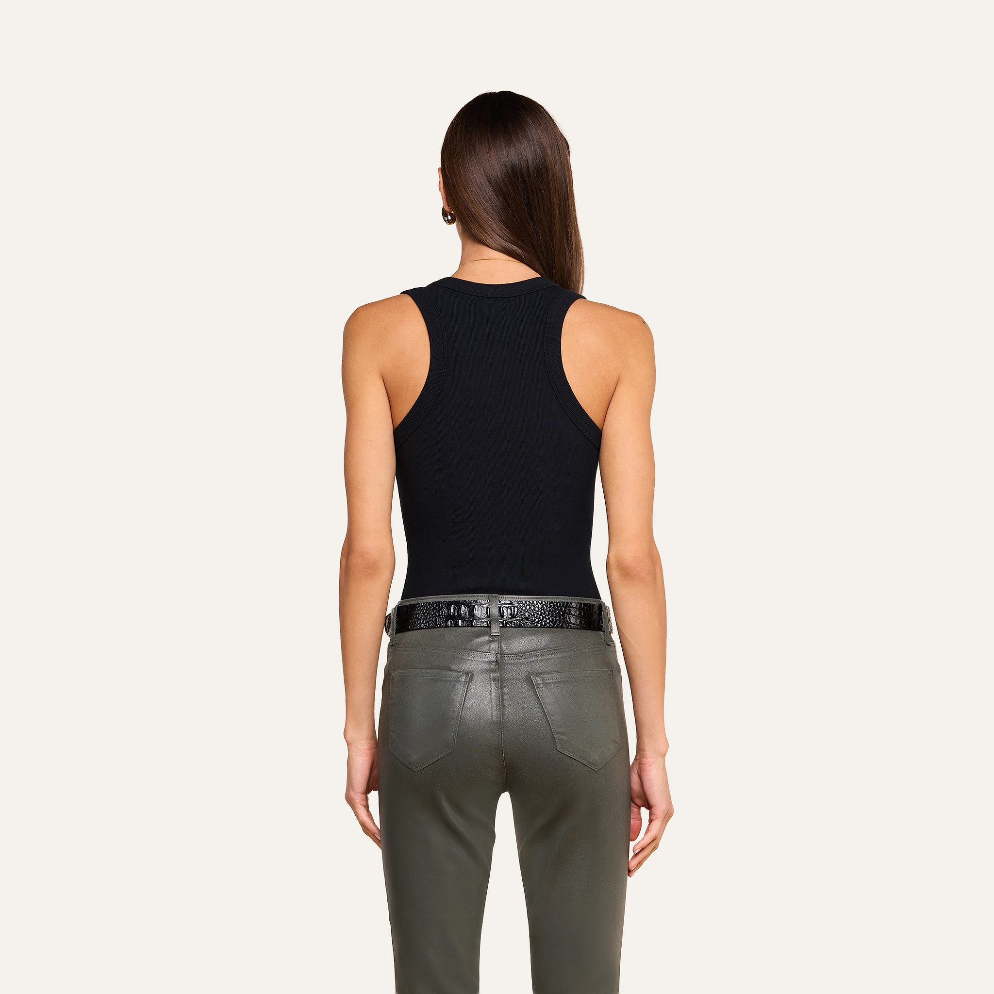 A person with long brown hair faces away, wearing the Shania Racerback Tank paired with a black belt and olive green leather pants against a plain beige background.