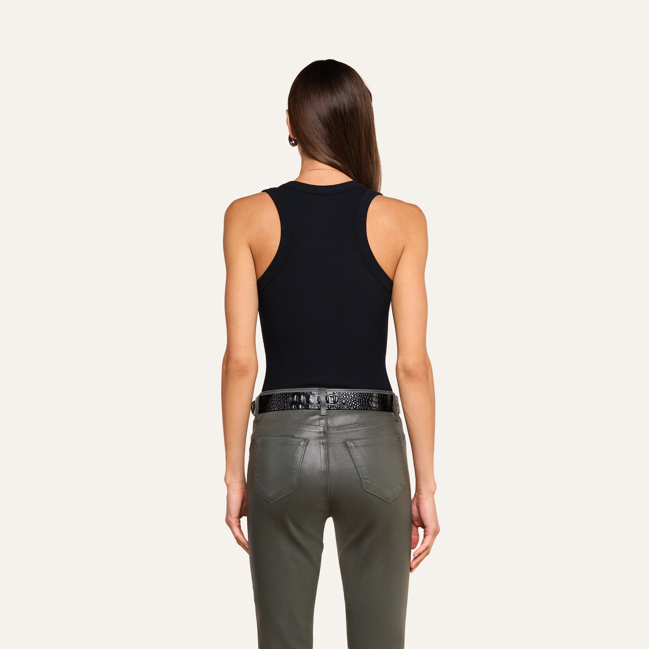 A person with long brown hair faces away, wearing the Shania Racerback Tank paired with a black belt and olive green leather pants against a plain beige background.