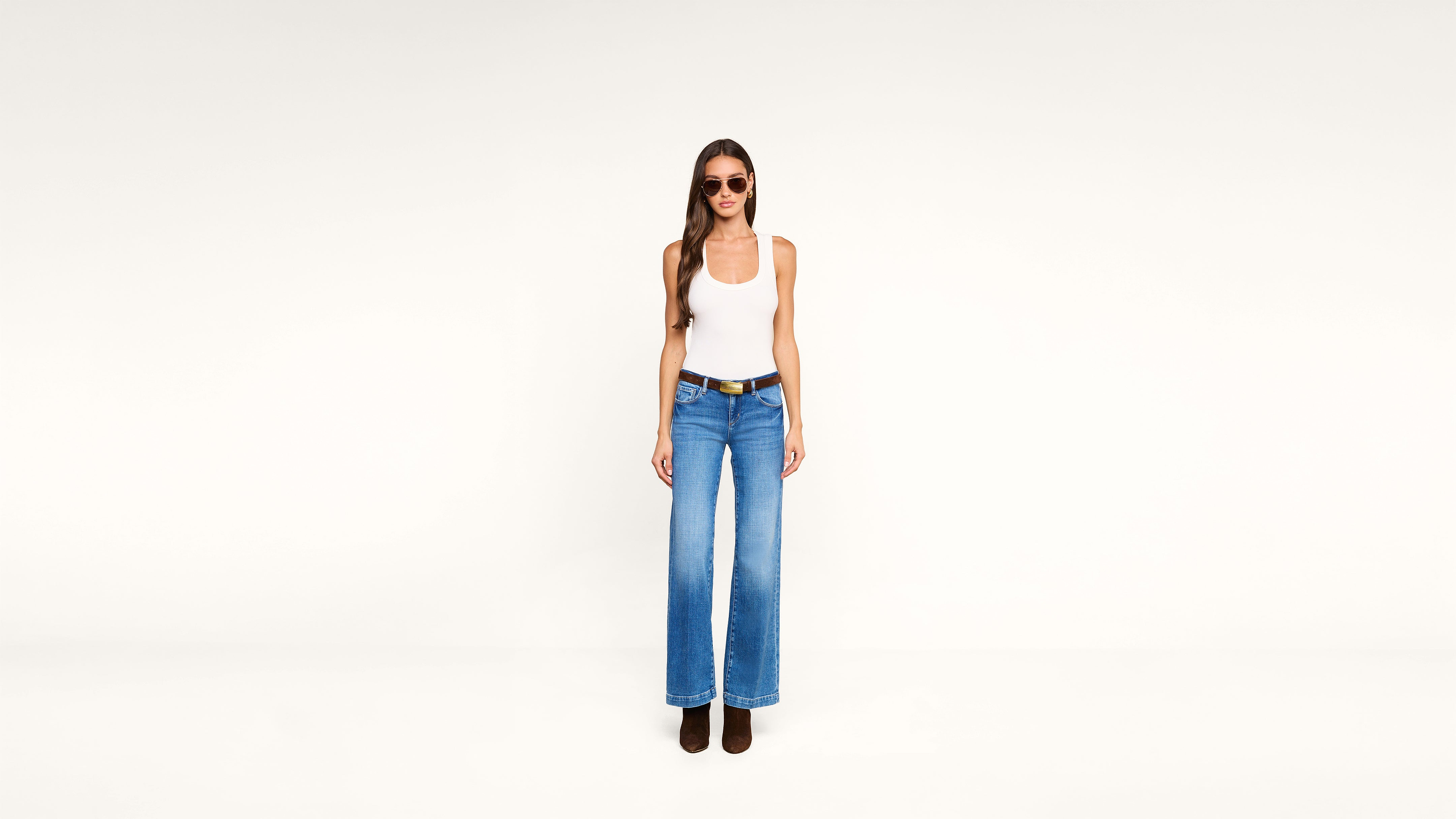 A woman wearing sunglasses, a white tank top, blue wide-leg jeans, and brown boots stands against a plain white background.