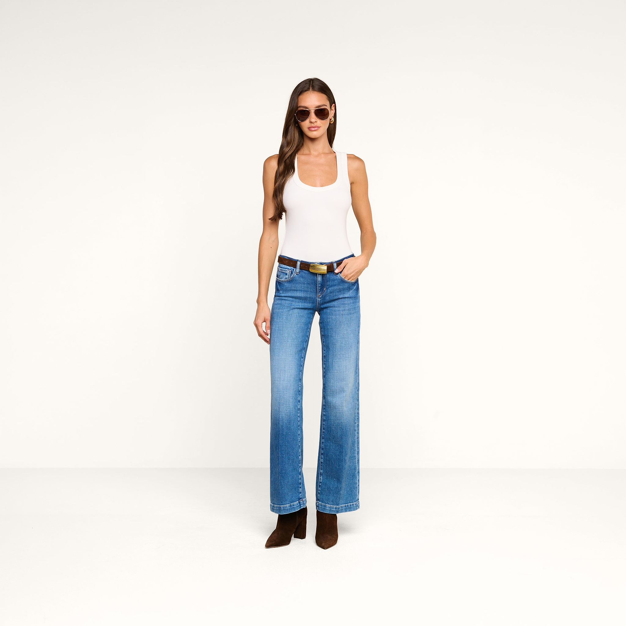 Una donna con gli occhiali da sole indossa la canotta Shania Racerback, jeans blu con una cintura nera e stivali marroni; è in piedi davanti a uno sfondo beige a tinta unita con una mano in tasca.