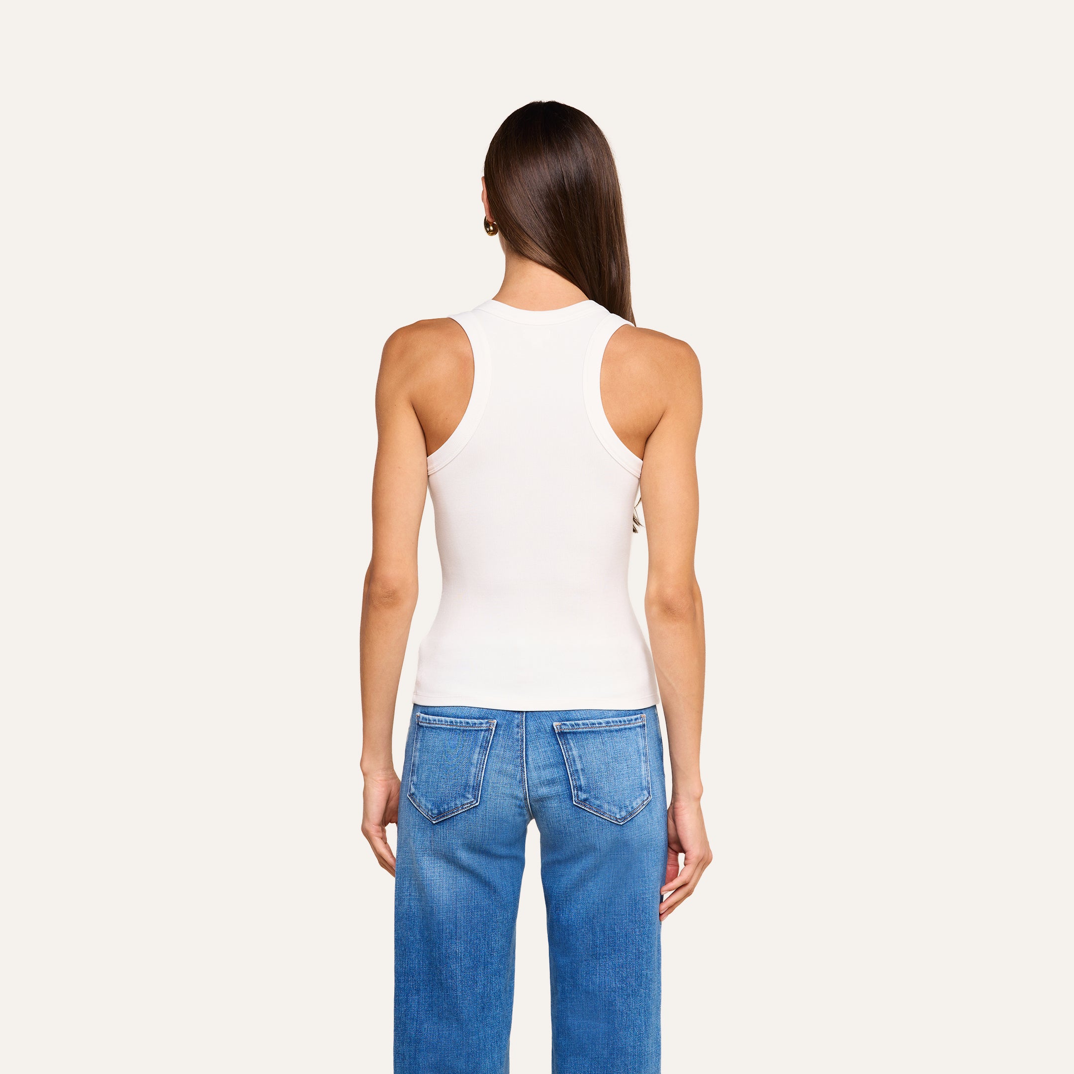 Una persona dai lunghi capelli castani, che indossa la canotta Shania Racerback in bianco e dei jeans blu, è in piedi di spalle alla fotocamera su uno sfondo beige a tinta unita.