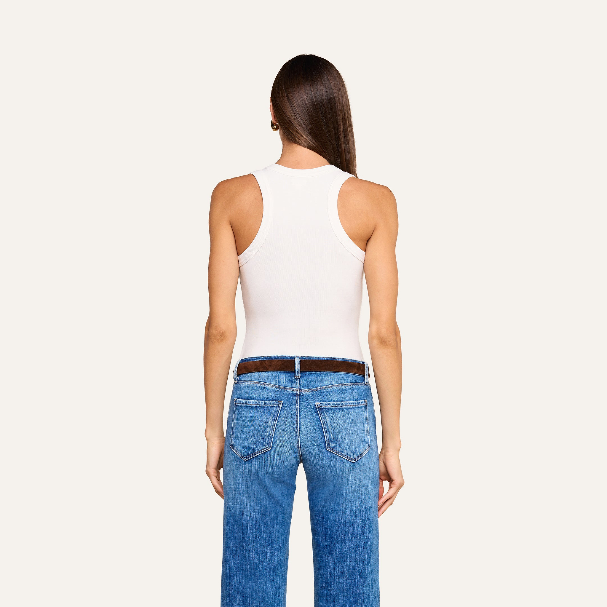 Una persona con lunghi capelli castani, che indossa la canotta Shania Racerback in bianco e jeans blu, è ritratta di schiena su uno sfondo beige a tinta unita.