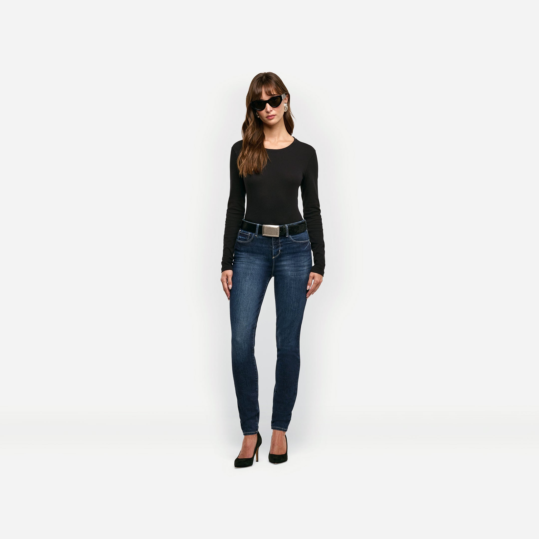 A woman wears a black Leanne Cotton Tee tucked into dark wash skinny jeans with a black belt and black high heels.
