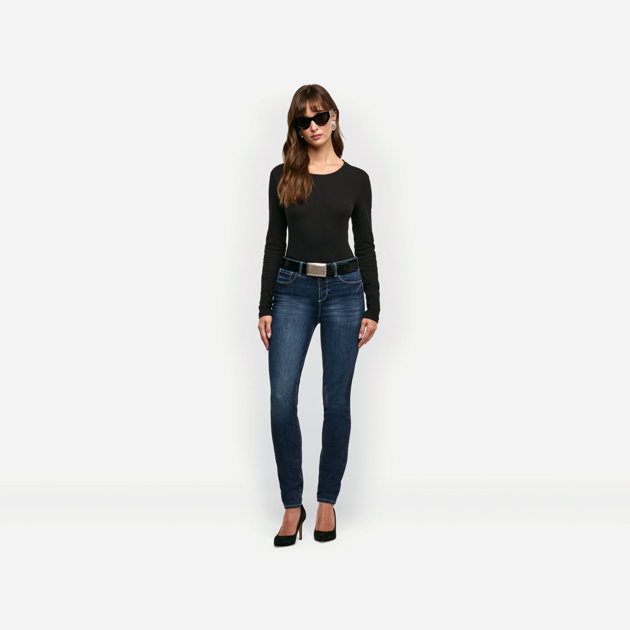 A woman wears a black Leanne Cotton Tee tucked into dark wash skinny jeans with a black belt and black high heels.