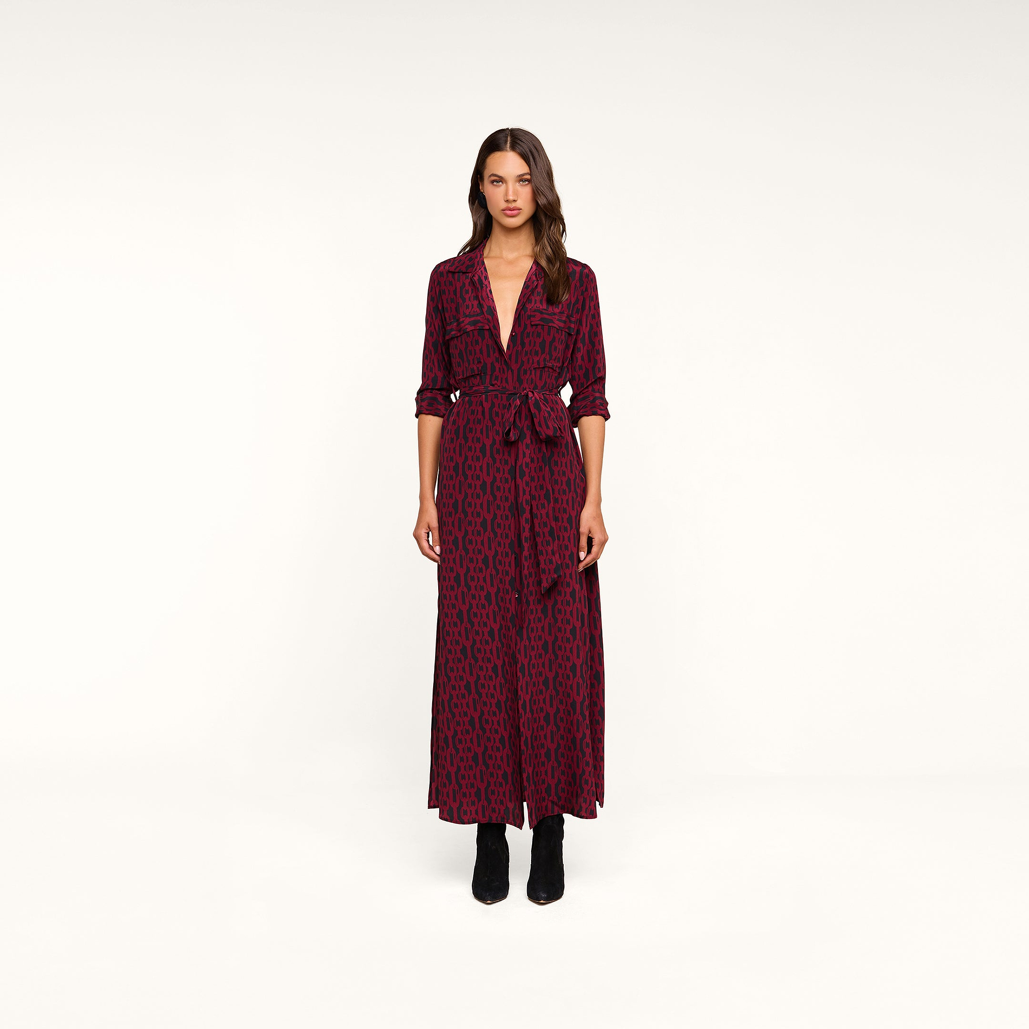 A woman stands in a studio wearing the Cameron Silk Shirt Dress—a long-sleeve, deep V-neck maxi shirtdress in maroon and black pattern with a tied waist—styled with black boots.