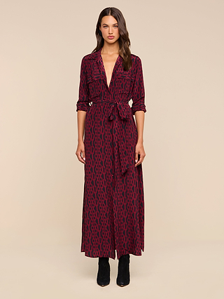 A woman stands in a studio wearing the Cameron Silk Shirt Dress—a long-sleeve, deep V-neck maxi shirtdress in maroon and black pattern with a tied waist—styled with black boots.