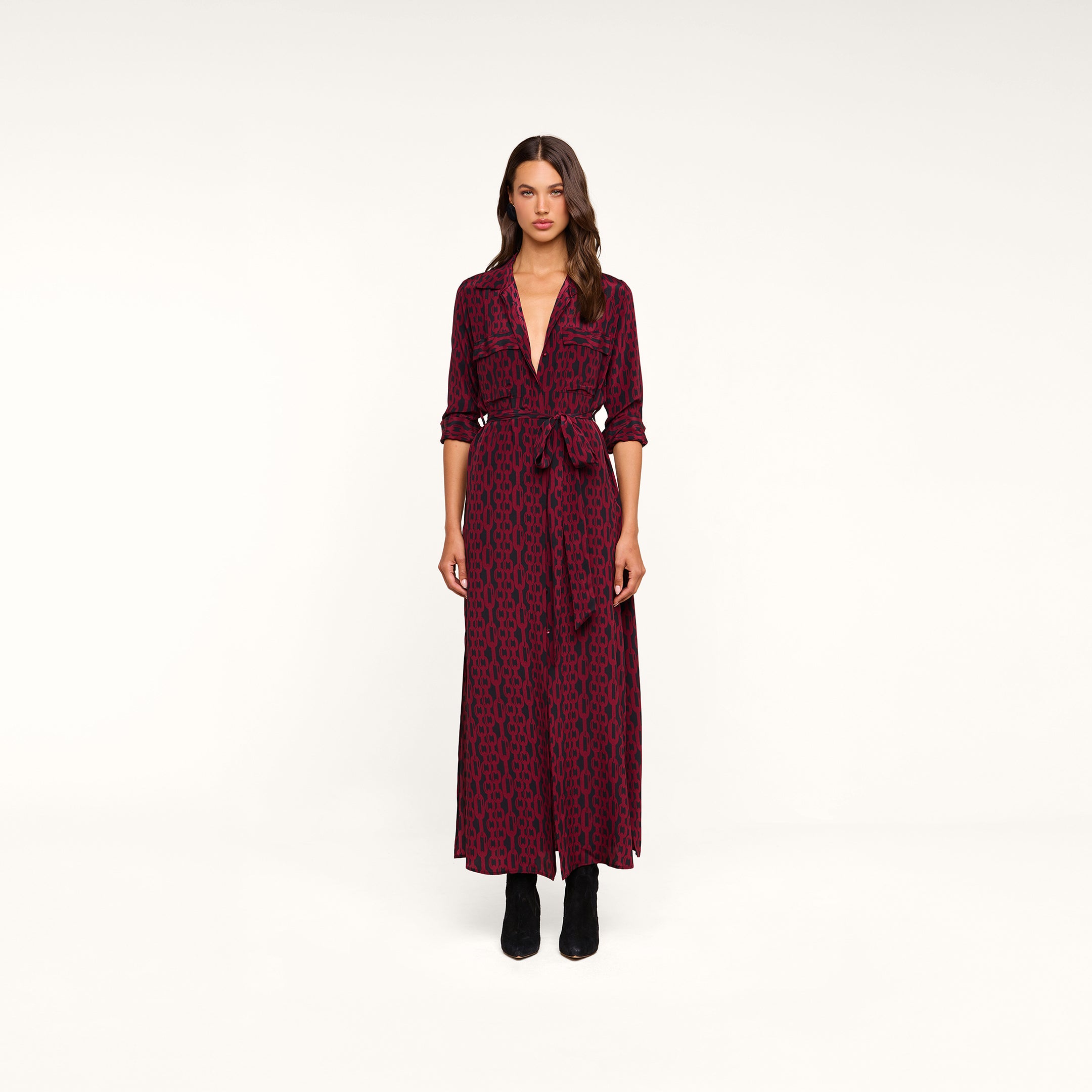A woman stands in a studio wearing the Cameron Silk Shirt Dress—a long-sleeve, deep V-neck maxi shirtdress in maroon and black pattern with a tied waist—styled with black boots.