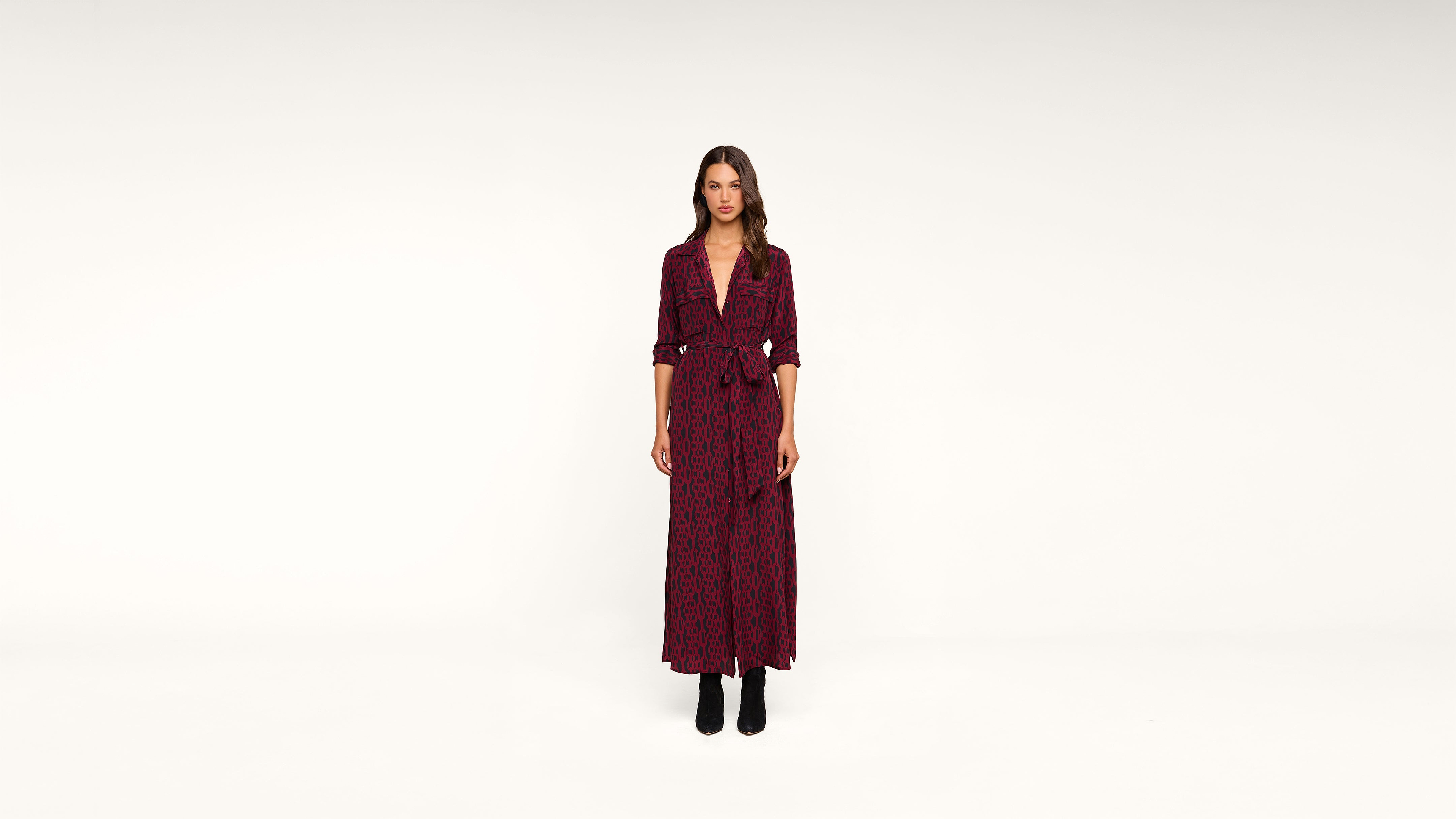 A woman stands against a plain white background, wearing a long, burgundy patterned dress with a V-neck and black ankle boots. Her hands are by her sides, and she looks directly at the camera.