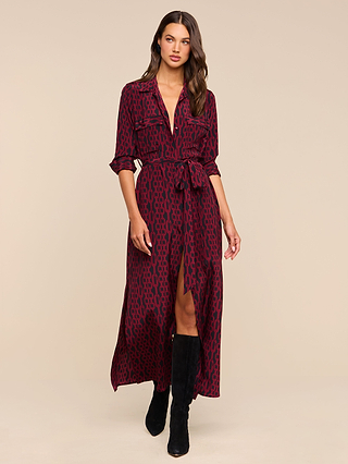 A woman stands against a plain beige background, wearing the Cameron Silk Shirt Dress—a long-sleeve, burgundy silk shirtdress with a black pattern and waist tie—paired with black knee-high boots.