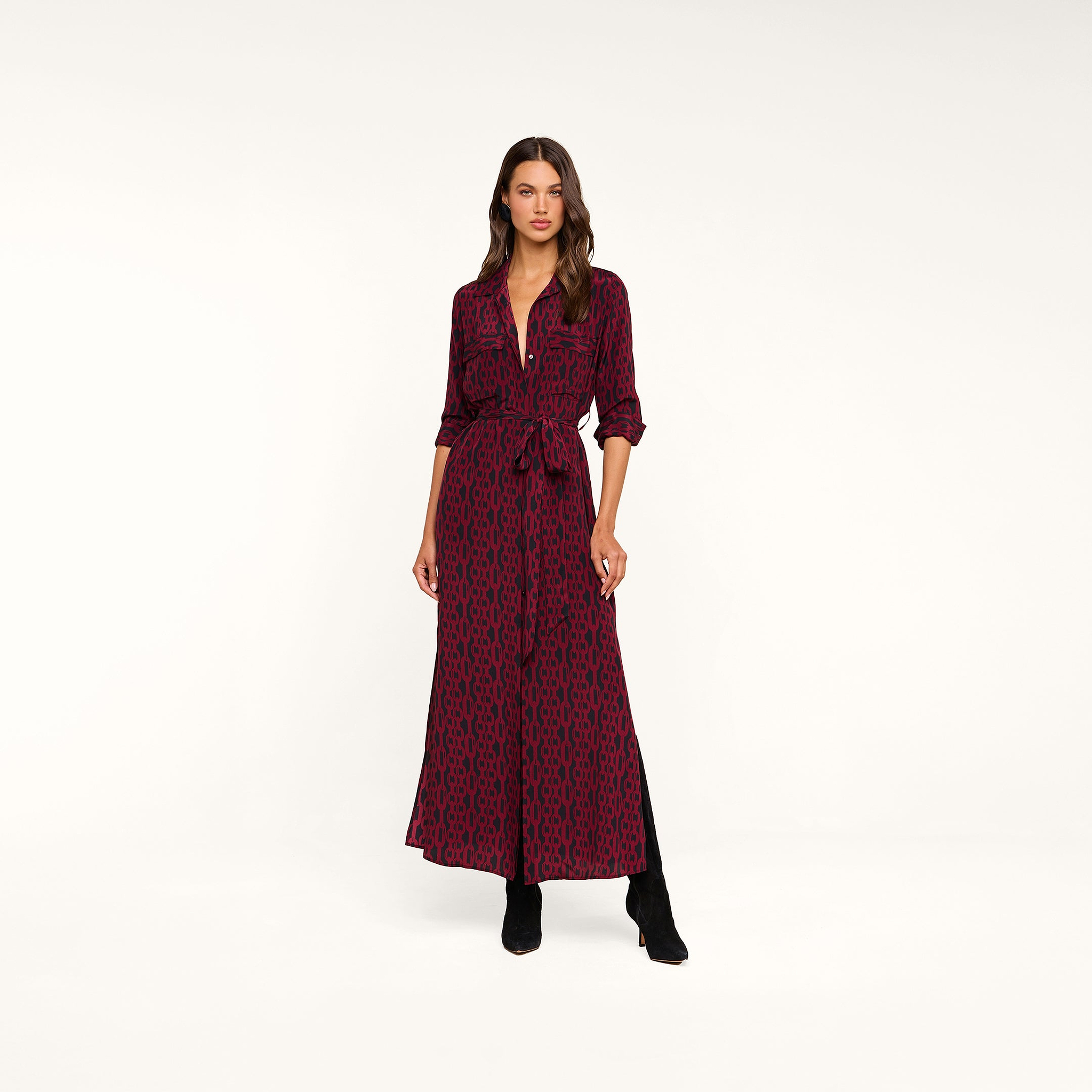 A woman stands against a plain beige background wearing the Cameron Silk Shirt Dress, a long-sleeve, button-down style in maroon with a black geometric pattern, cinched at the waist and paired with black boots.