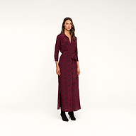 A woman stands against a plain beige background wearing the Cameron Silk Shirt Dress, featuring long sleeves, ankle-length maroon silk fabric with a black geometric pattern, a tied waist belt, and black boots.