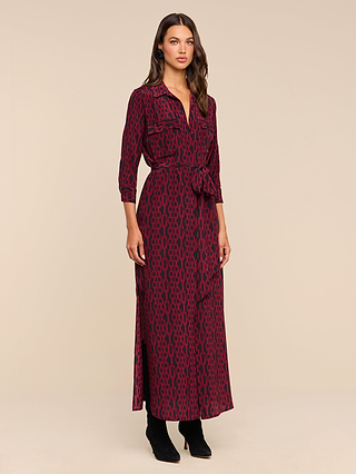 A woman stands against a plain beige background wearing the Cameron Silk Shirt Dress, featuring long sleeves, ankle-length maroon silk fabric with a black geometric pattern, a tied waist belt, and black boots.