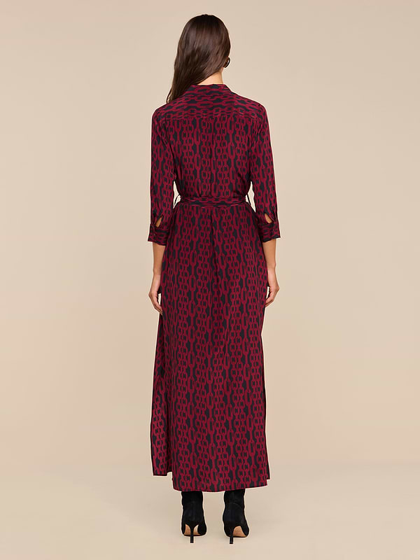 A woman stands facing away, wearing the Cameron Silk Shirt Dress—a maroon maxi dress with a black geometric pattern and three-quarter sleeves—paired with black heeled boots against a plain beige background.