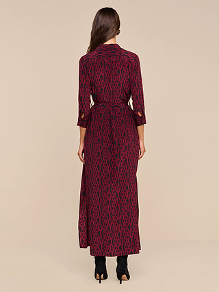A woman stands facing away, wearing the Cameron Silk Shirt Dress—a maroon maxi dress with a black geometric pattern and three-quarter sleeves—paired with black heeled boots against a plain beige background.