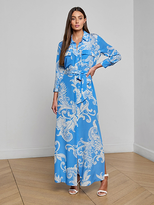 A woman stands indoors in the Cameron Silk Shirt Dress, a long blue silk piece with a white paisley print, belted waist, button front, and long sleeves. She has long brown hair and pairs the dress with white heeled sandals.