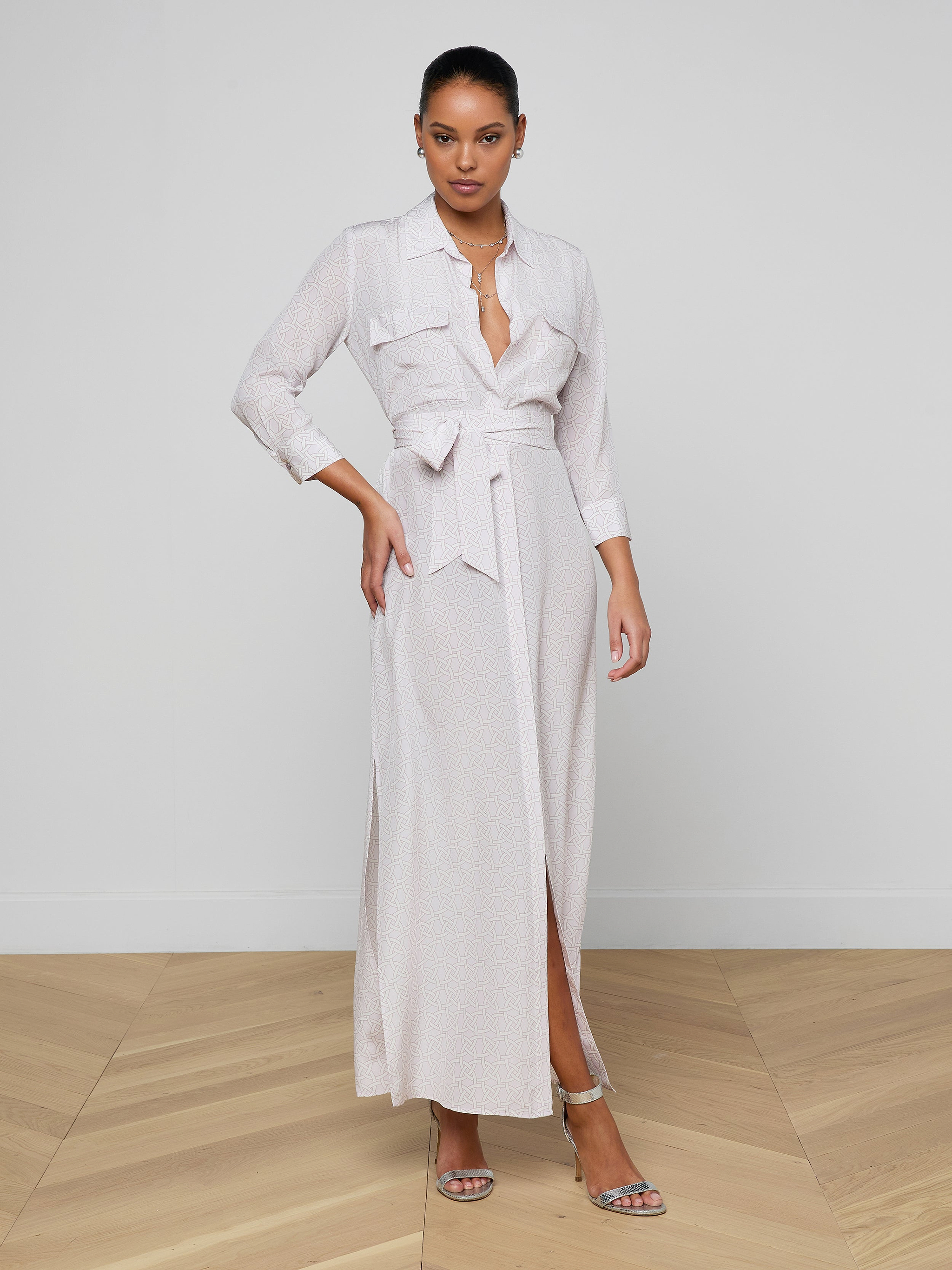 A woman stands indoors on a wooden floor, confidently posing in the Cameron Silk Shirt Dress—a long, light-colored silk dress with a deep neckline and long sleeves. Her hair is tied back and she completes the look with sandals.