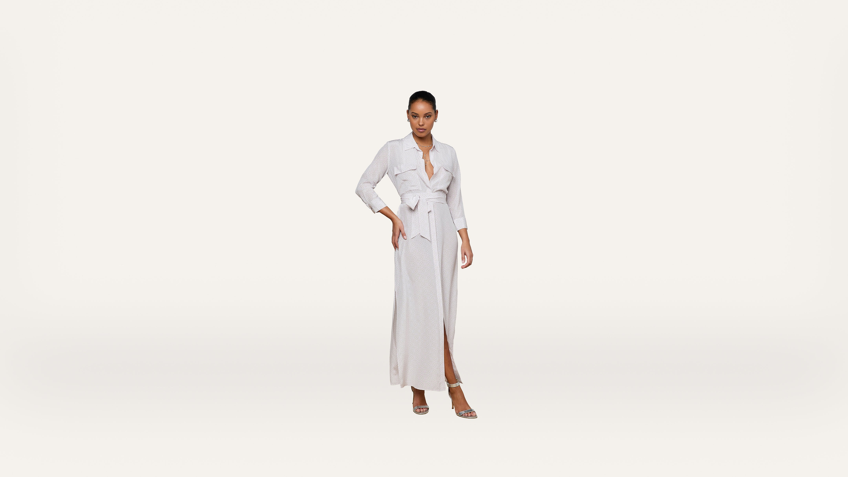 A woman stands confidently wearing a light-colored, long-sleeve wrap dress with a waist tie and slit, paired with heeled sandals, against a plain white background.