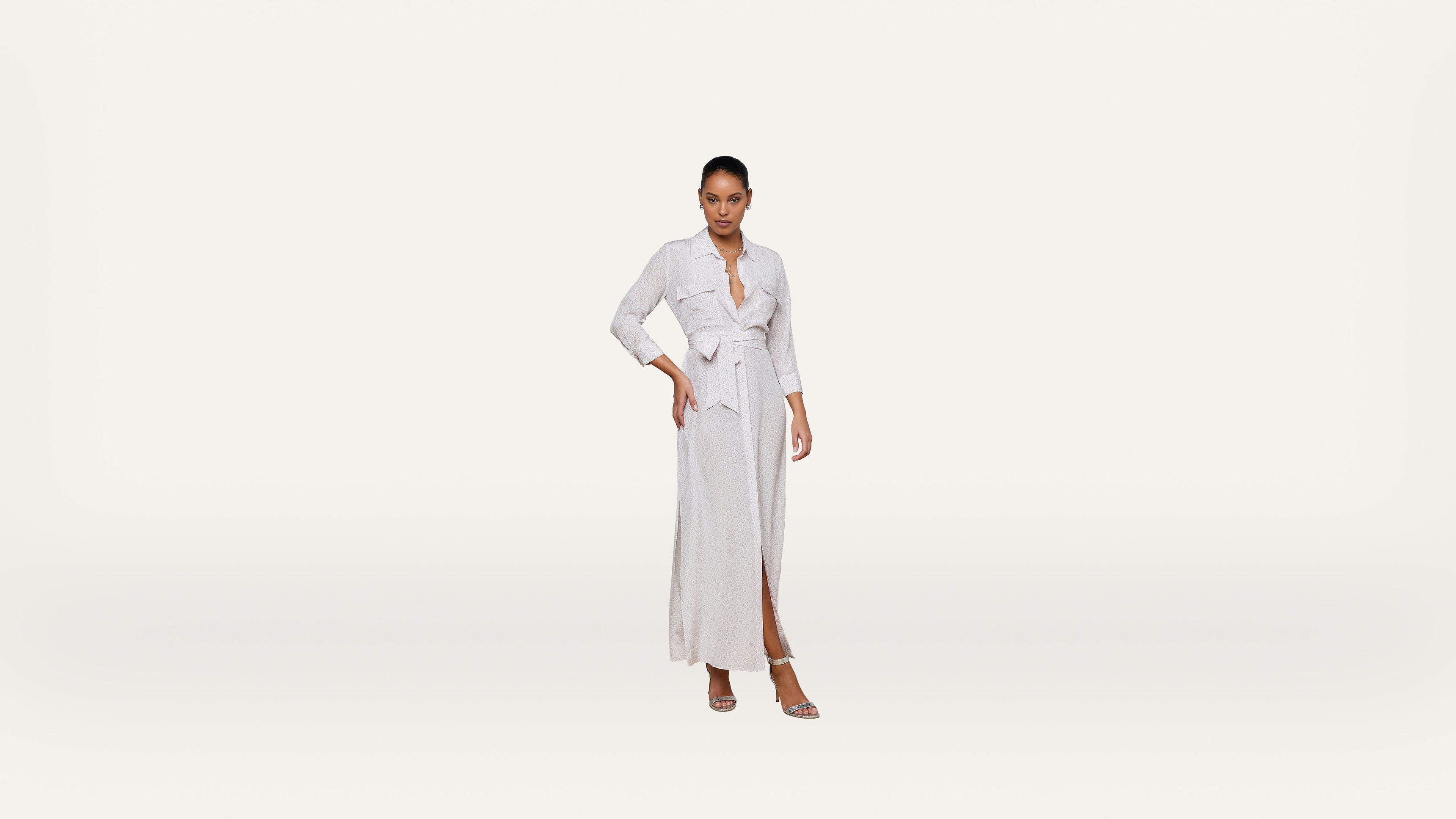 A woman stands confidently wearing a light-colored, long-sleeve wrap dress with a waist tie and slit, paired with heeled sandals, against a plain white background.