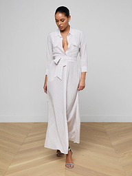 A woman stands on a wooden floor in the Cameron Silk Shirt Dress, a long, light-colored silk crepe de chine dress with a deep V-neck, long sleeves, and waist tie. Her hair is pulled back as she looks down, wearing strappy heeled sandals.